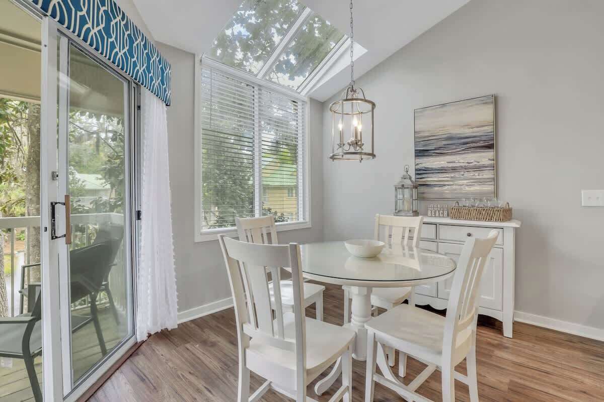 A dining area with a glass table, white chairs, a chandelier, and a view of the outdoors through large windows.