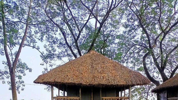 Eine Reetdachhütte mit Holzbalkon, umgeben von Bambuszaun und Grünpflanzen.