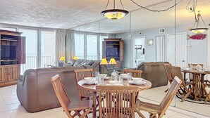 A spacious living area with a large sofa, a dining table set for four, and a television mounted on the wall.