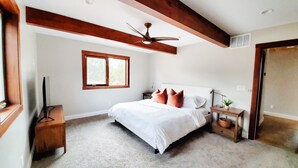 Un dormitorio con cama, cabecera de madera, ventilador de techo, ventana, mesita de noche, lámpara y una planta pequeña.