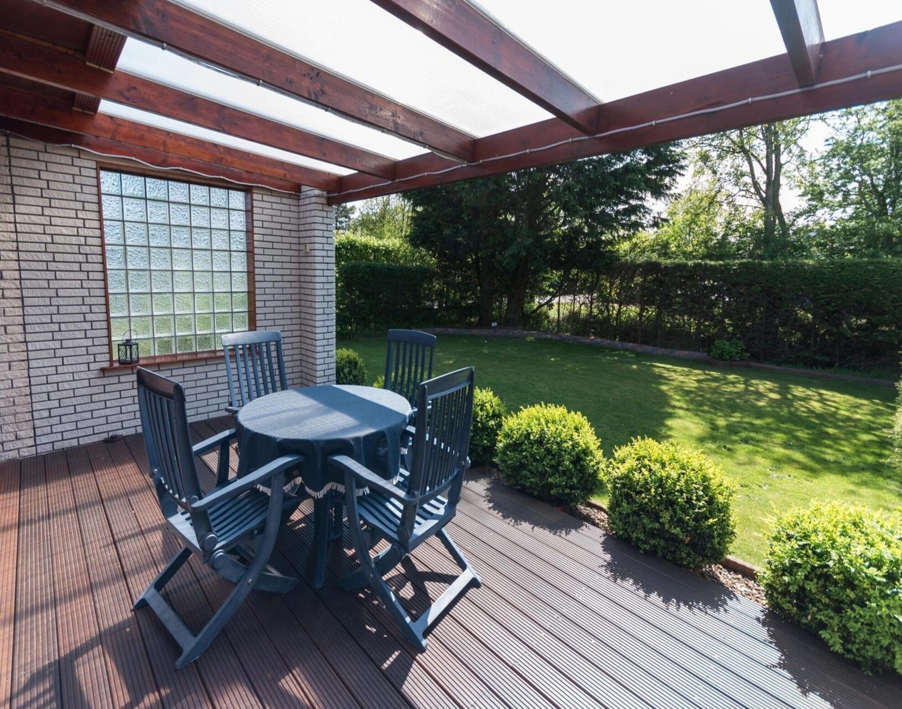 A covered patio with a wooden deck, a round table with chairs, and a brick wall with a window.