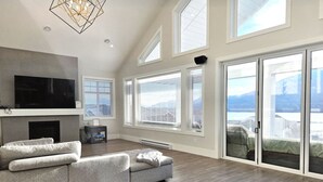 A spacious living room with a large sectional sofa, a flat-screen TV mounted on the wall, and a fireplace. There are large windows and a sliding glass door leading to a balcony. - Sky-View Cottage - 5 Bedroom View home 265 (Osoyoos)