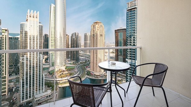 A balcony with two wicker chairs and a small round table, overlooking a cityscape with high-rise buildings and a river. - Lake View Studio near Marina with Pool, Sauna and Gym (Dubai)