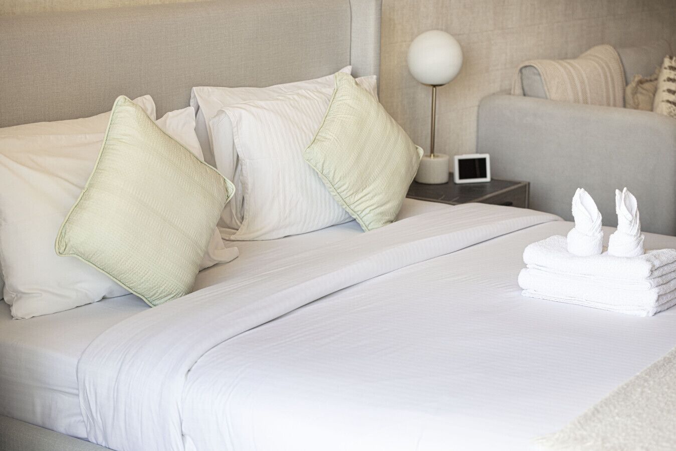 Een netjes opgemaakt bed met witte lakens en kussens, een nachtkastje met een lamp en een klein elektronisch apparaat, en een stapel opgevouwen handdoeken.