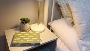 A neatly made bed with a checkered pillow and a floral blanket. A white bedside table holds a lamp, a small potted plant, and a book titled 'Pernicious Jane Austen'. - Creeslough - 2 Bed Cottage - Pet Friendly - Garden (Glenveagh)