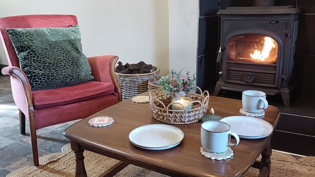 A cozy living room with a red armchair, a wooden coffee table with plates and cups, a fireplace, and a wicker basket with decorative items. - Creeslough - 2 Bed Cottage - Pet Friendly - Garden (Glenveagh)