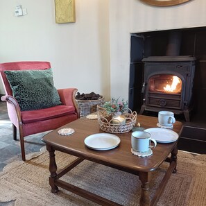 A cozy living room with a red armchair, a wooden coffee table with plates and cups, a fireplace, and a wicker basket with decorative items. - Creeslough - 2 Bed Cottage - Pet Friendly - Garden (Glenveagh)