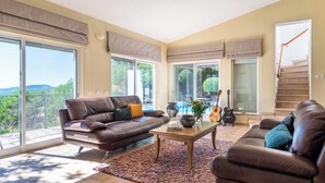 Un salon spacieux avec un canapé en cuir marron, une table basse et un tapis coloré. De grandes fenêtres avec des rideaux, un escalier et une vue sur l’extérieur complètent l’ensemble.
