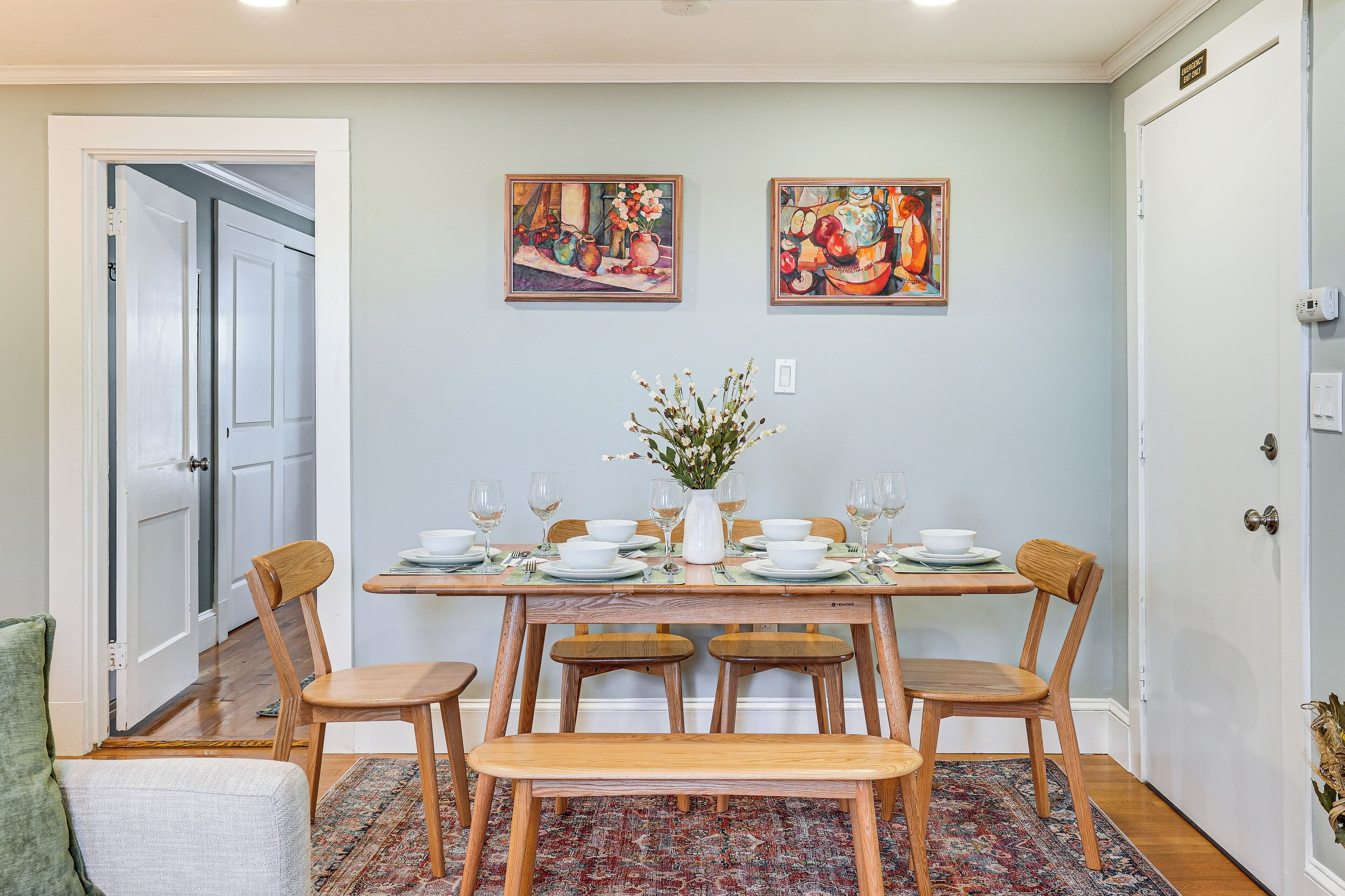 A dining area with a rectangular table set for six, surrounded by wooden chairs and benches. There are two framed artworks on the wall above the table.