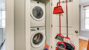 A laundry room with a front-loading washer and dryer, a red broom, a dustpan, and a bucket. - Walk to Constitution Beach: Mid-century Boston Gem (Boston)
