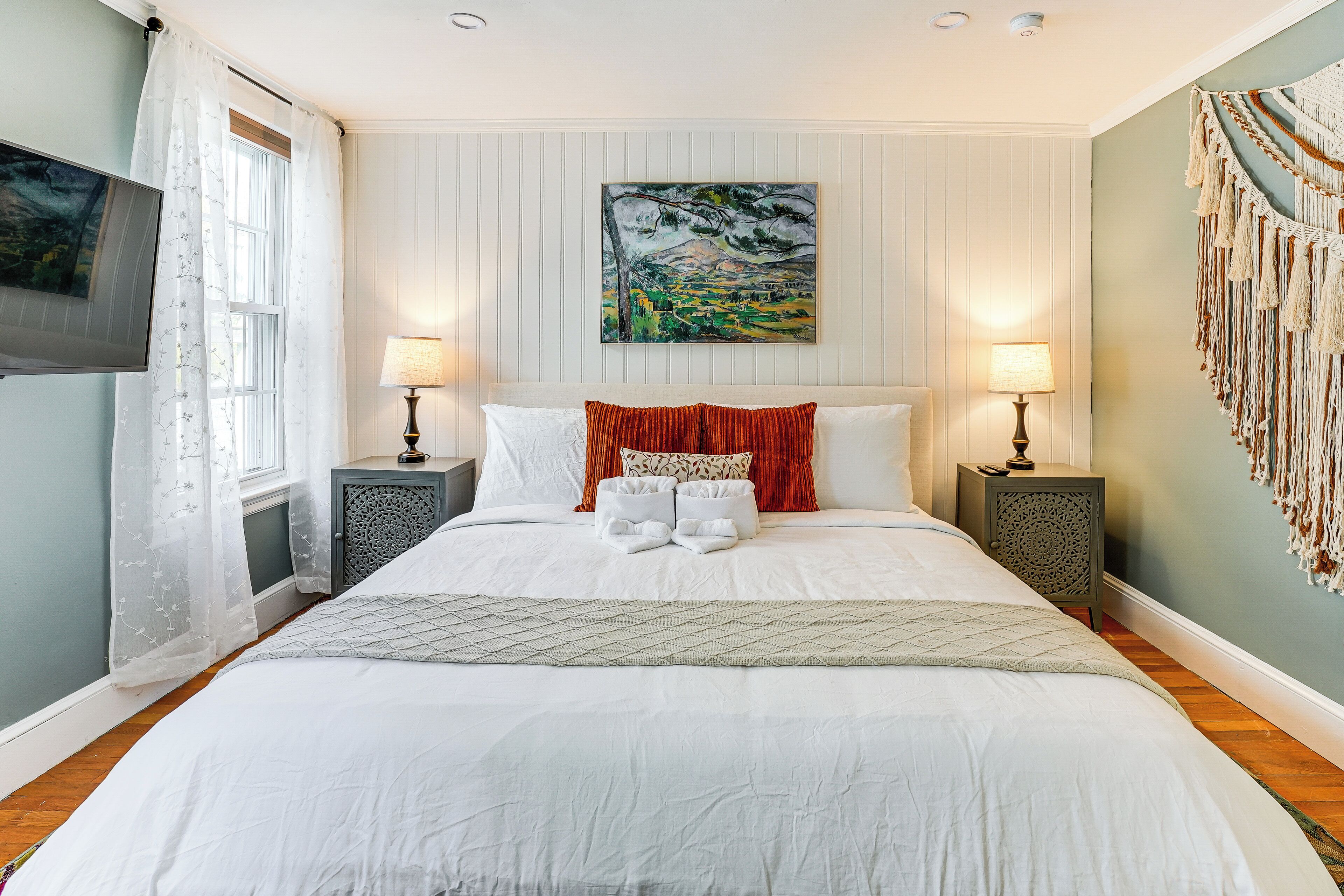 A neatly made bed with white linens, two bedside tables with lamps, a painting above the bed, and a macrame wall hanging.