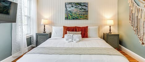 A neatly made bed with white linens, two bedside tables with lamps, a painting above the bed, and a macrame wall hanging.