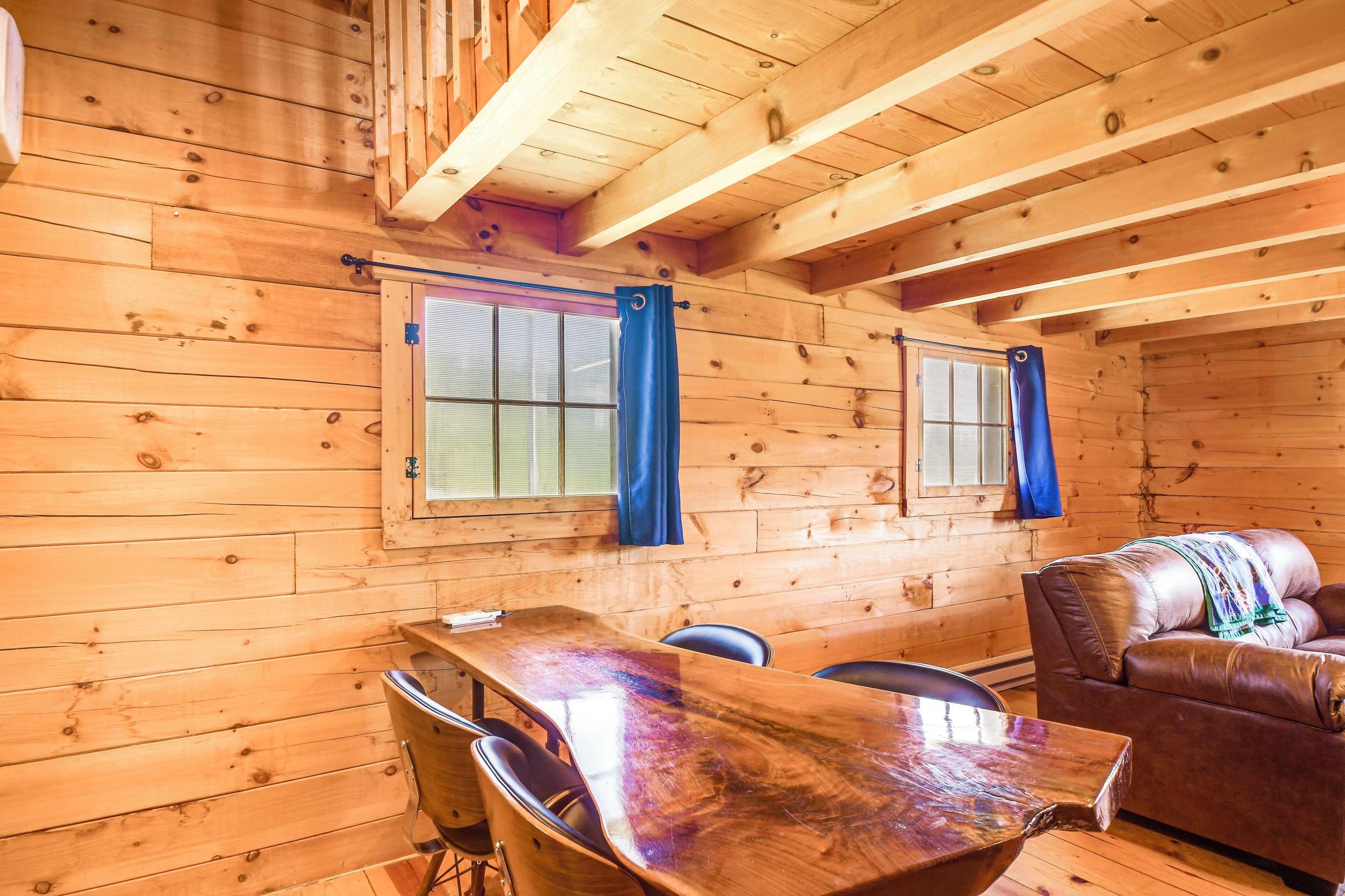 A wooden cabin interior with a dining table, chairs, a sofa, and two windows with curtains.