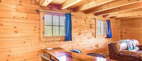 A wooden cabin interior with a dining table, chairs, a sofa, and two windows with curtains.