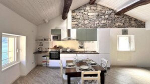 A kitchen with a stone wall, wooden beams, a dining table set for four, and a view of the sea through a window. - Casa Hygge Argegno (Argegno)