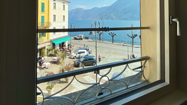 A view from a window overlooking a street with buildings, parked cars, and a body of water with mountains in the background. - Casa Hygge Argegno (Argegno)