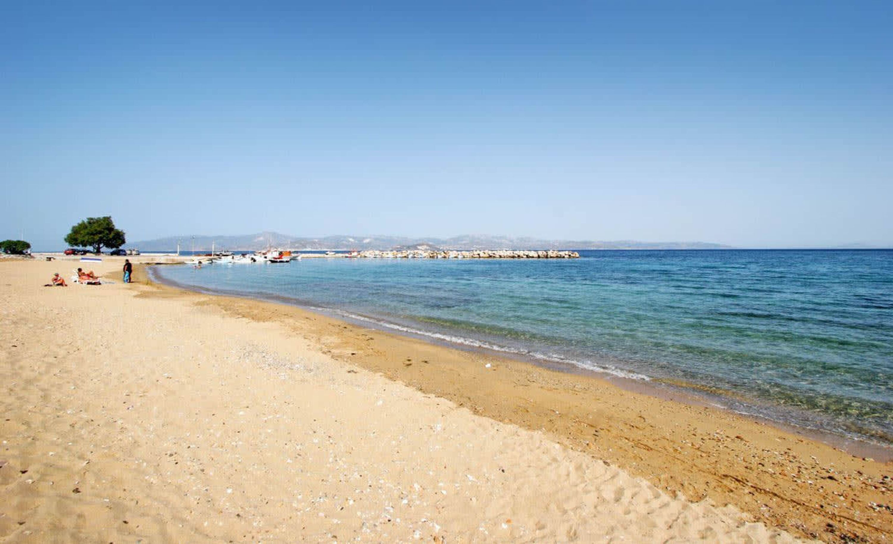 A beach with clear skies, calm waters, and a rocky breakwater in the distance.