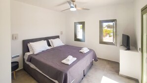 A modern bedroom with a large bed, a ceiling fan, two windows, and a TV on a stand. - Ampelas Nostalgia Apartments - Aiolos Gem in Paros (Paros)