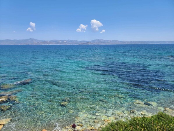 A coastal view with clear turquoise waters, rocky shoreline, and distant mountains under a clear sky.