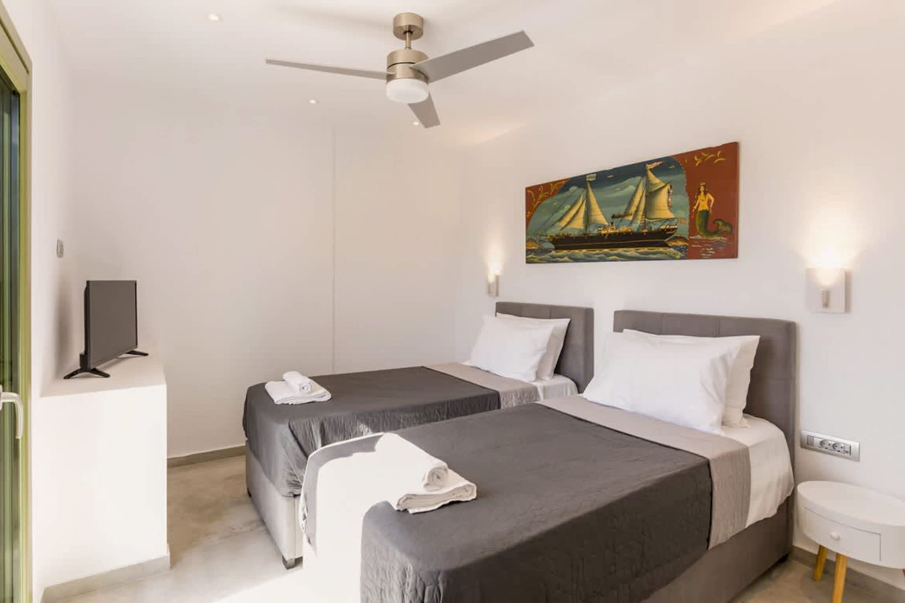 Ein Hotelzimmer mit zwei Betten, einem Deckenventilator, einem Bild eines Segelbootes und einem Fernseher auf einem Tisch.