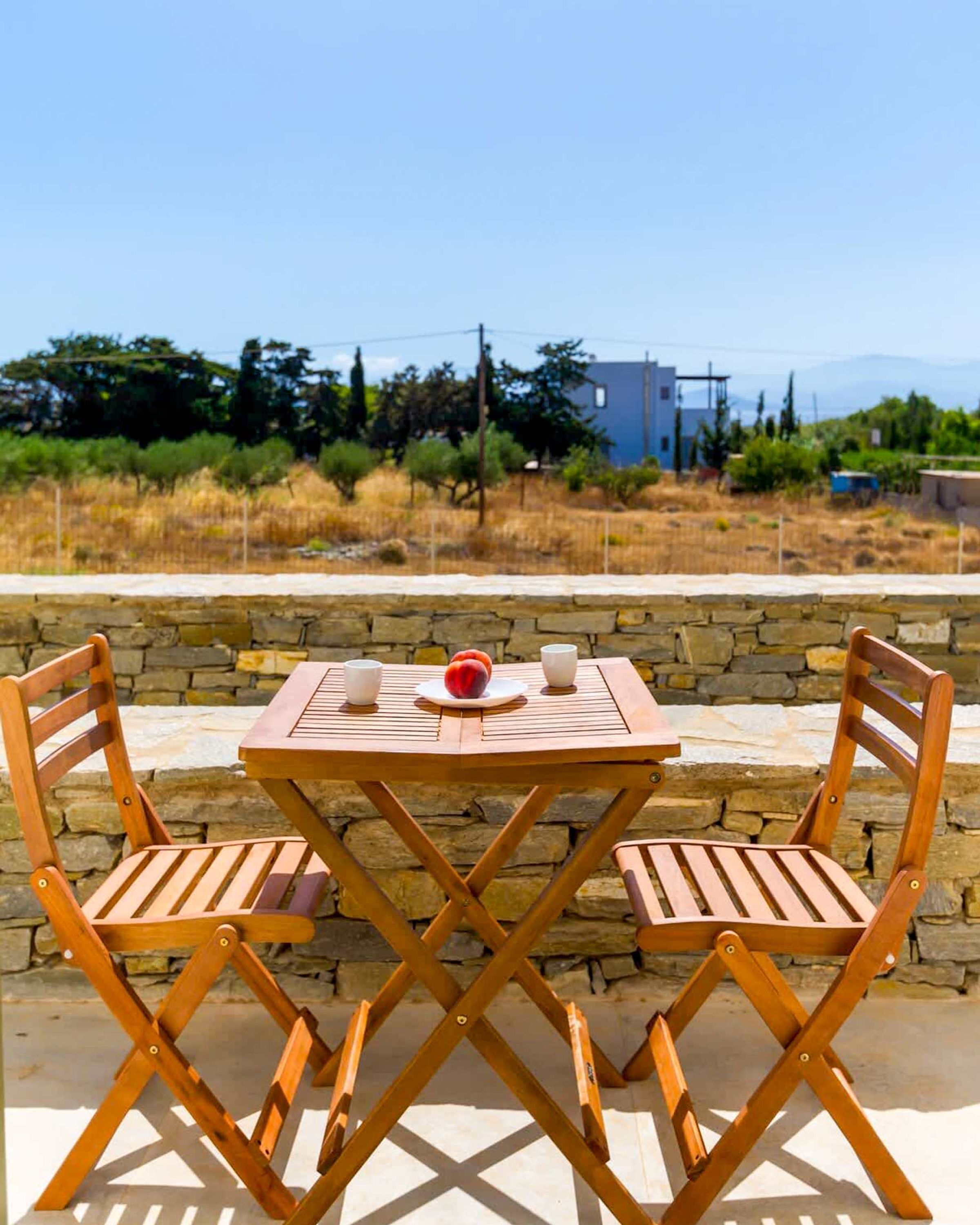 A wooden table and chairs set outdoors with a stone wall and a view of a building and trees in the background.