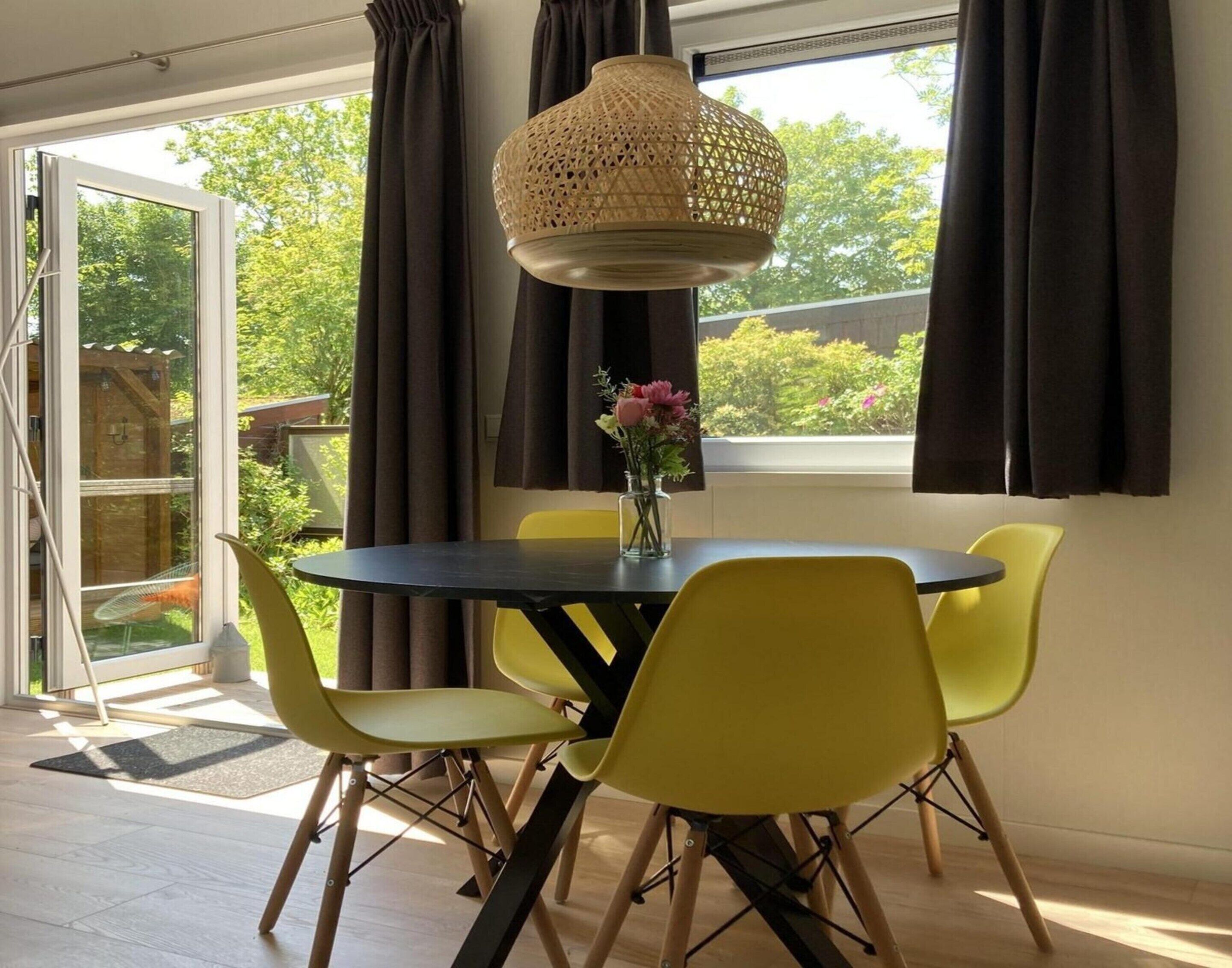 A dining area with a round table, four yellow chairs, a vase with flowers, and a woven pendant light.