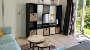 A living room with a sofa, two round wooden tables, a TV on a black entertainment unit, and a large window with curtains. - Modern Chalet With Garden, Terrace (Sint Maartensvlotbrug)