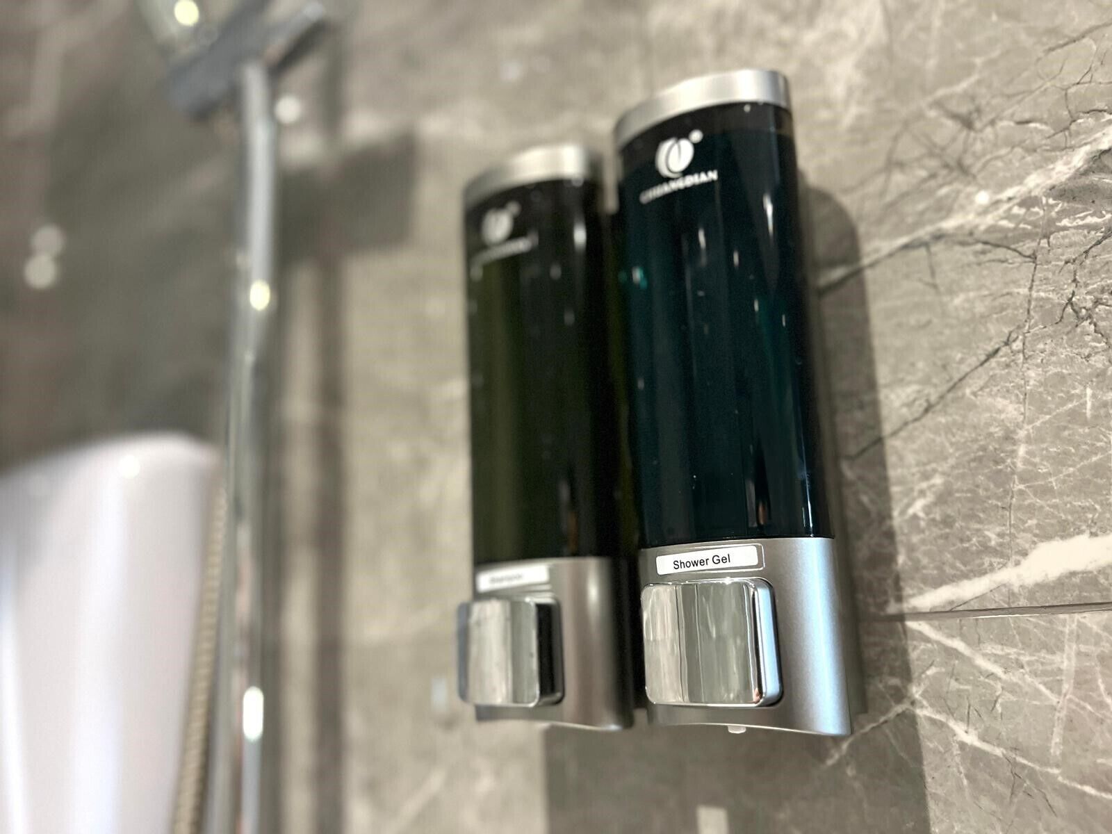 A bathroom with a marble wall, a shower, and two bottles labeled "Shower Gel" on a shelf.