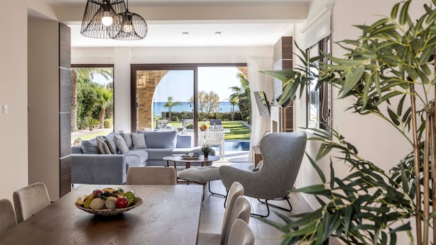 Ein modernes Wohnzimmer mit Esstisch, grauem Sofa und einem Sessel. Ein großes Fenster bietet Blick auf einen Pool und Palmen.