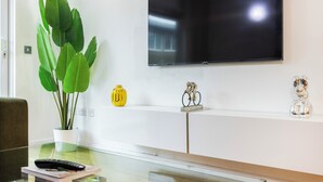 A modern living room with a flat-screen TV mounted on the wall, a glass coffee table, a potted plant, and decorative items on a shelf. - Grain Store One Bed Luxury Property  (London)