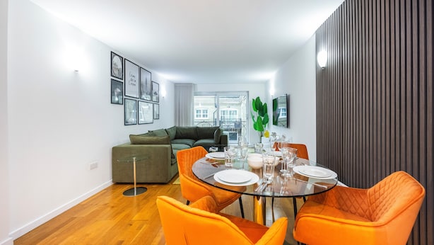 A modern living room with a glass dining table, orange chairs, and a green sofa. - Grain Store One Bed Luxury Property  (London)