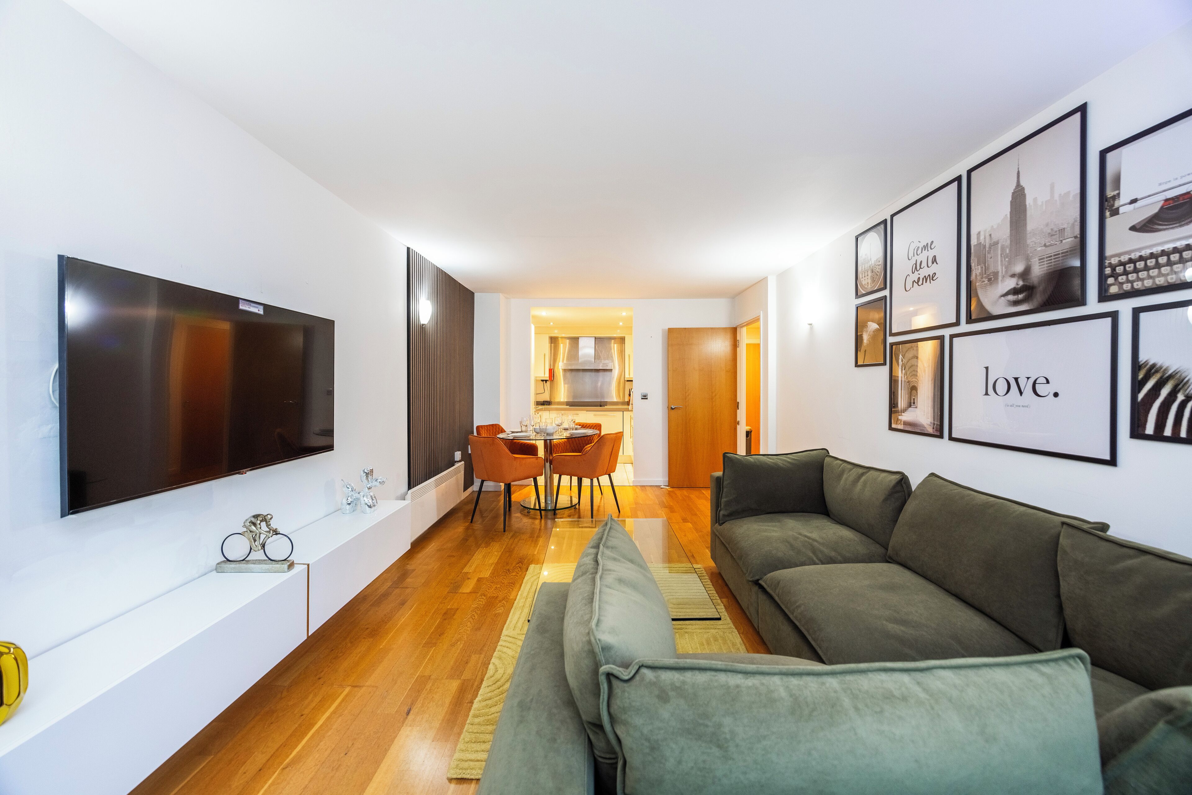 A modern living room with a flat-screen TV, a green sofa, and a dining area with orange chairs.