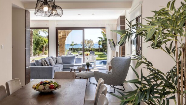 A modern living room with a dining area, a grey sofa, a round table with a fruit bowl, and a view of a pool and greenery. - Ayia Napa Seafront Ionion Villa H5 (Sotira)