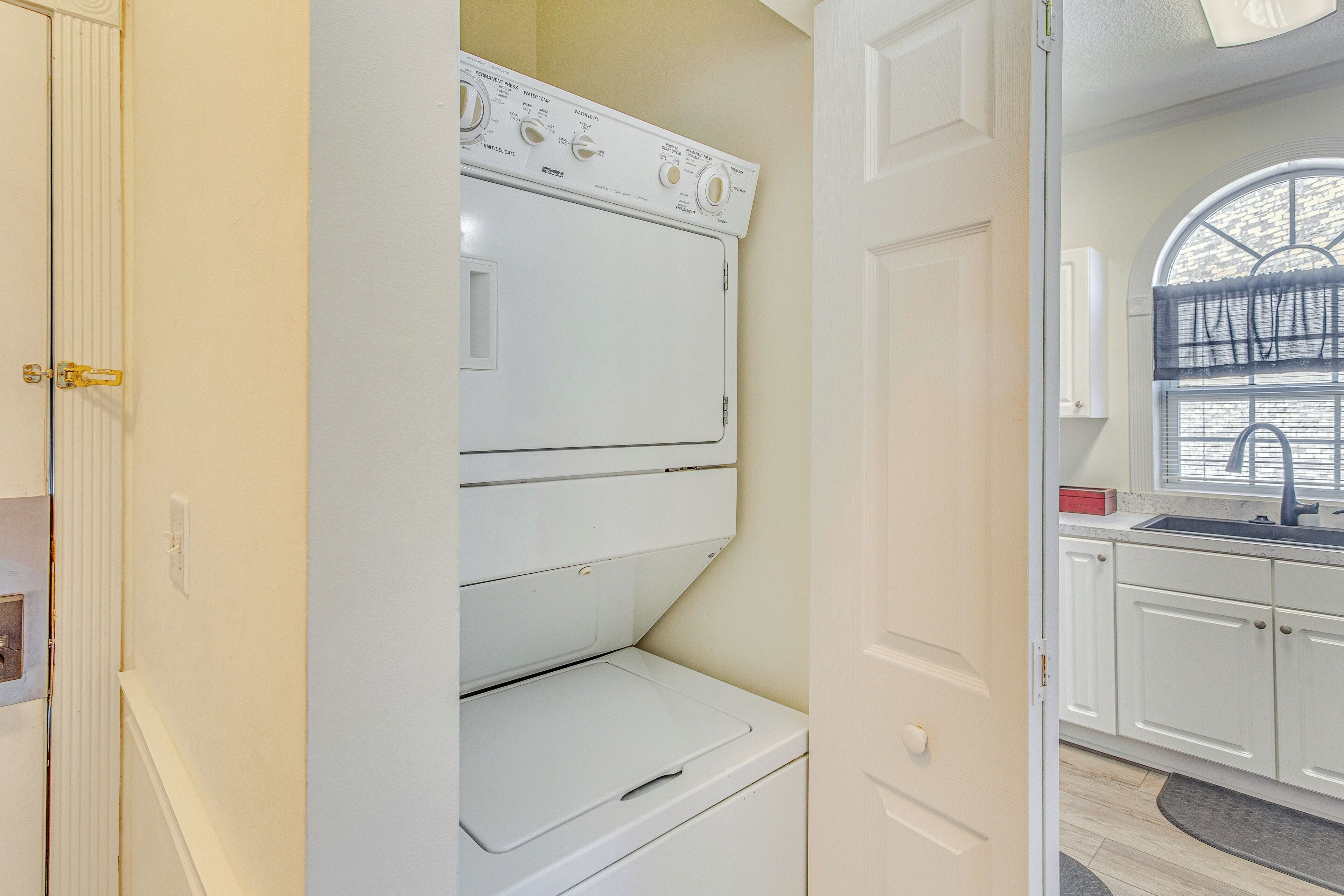 A laundry room with a stacked washer and dryer, a window with blinds, and a sink with a red soap dish.
