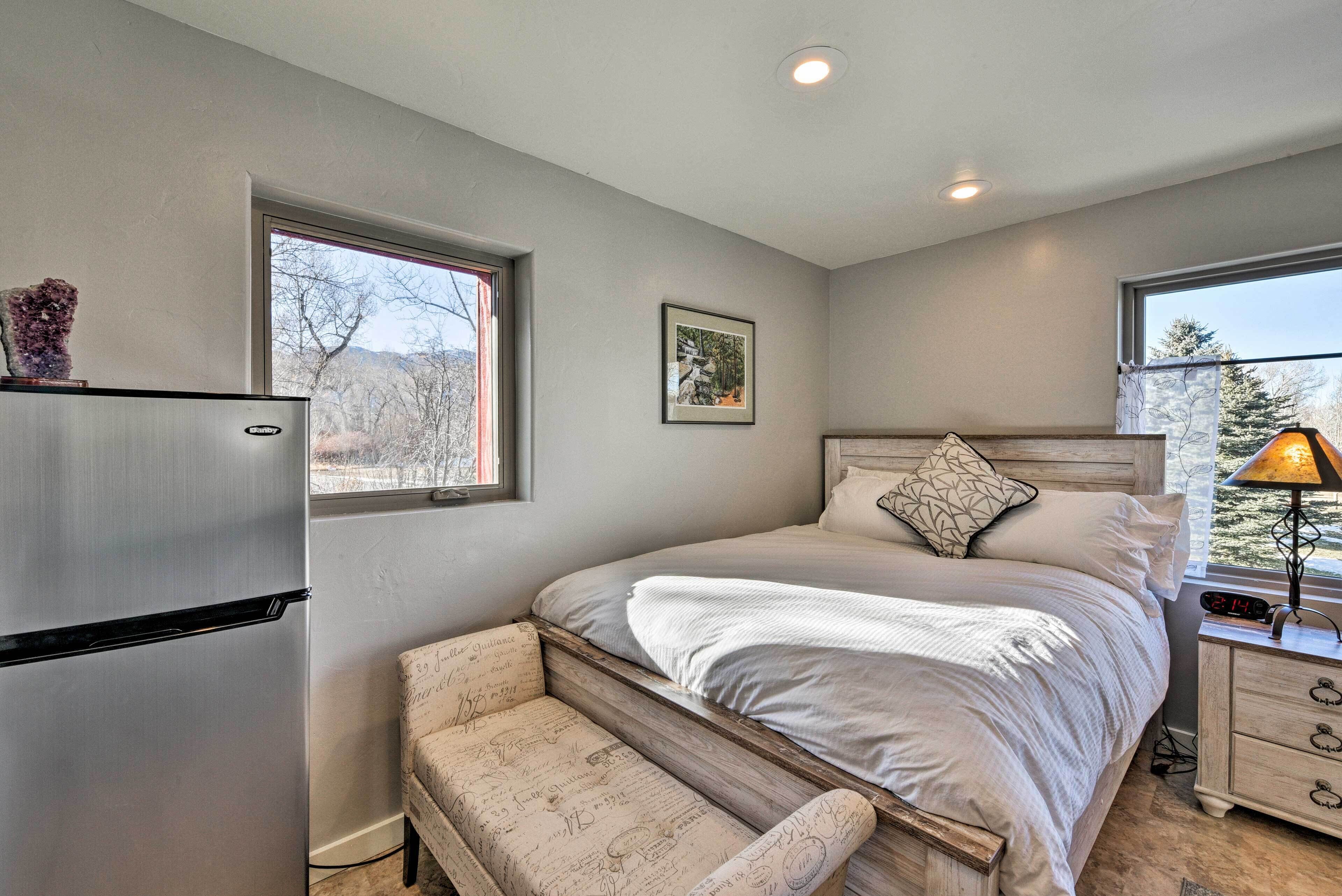 Un dormitorio con una cama, un banco, un refrigerador, una mesita de noche y una ventana.