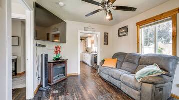 A living room with a dark wood floor, a gray sofa with an orange pillow, a ceiling fan, and a large window.