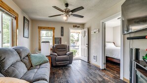 Ein Zimmer mit dunklem Holzboden, einem grauen Sofa, einem Liegestuhl, einem Deckenventilator und einem Bett mit weißer Matratze.