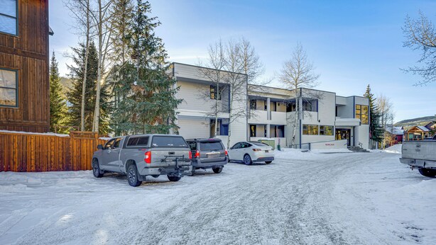 Ein Gebäude mit modernem Design, das Holz und Metall kombiniert. Vor dem Gebäude sind mehrere geparkte Fahrzeuge, und der Boden ist mit Schnee bedeckt.