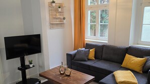 A modern living room with a flat-screen TV, a wooden coffee table, a grey sofa with yellow cushions, and a window with curtains. - WohnTraum zur alten Post, old town, sauna (Meißen)