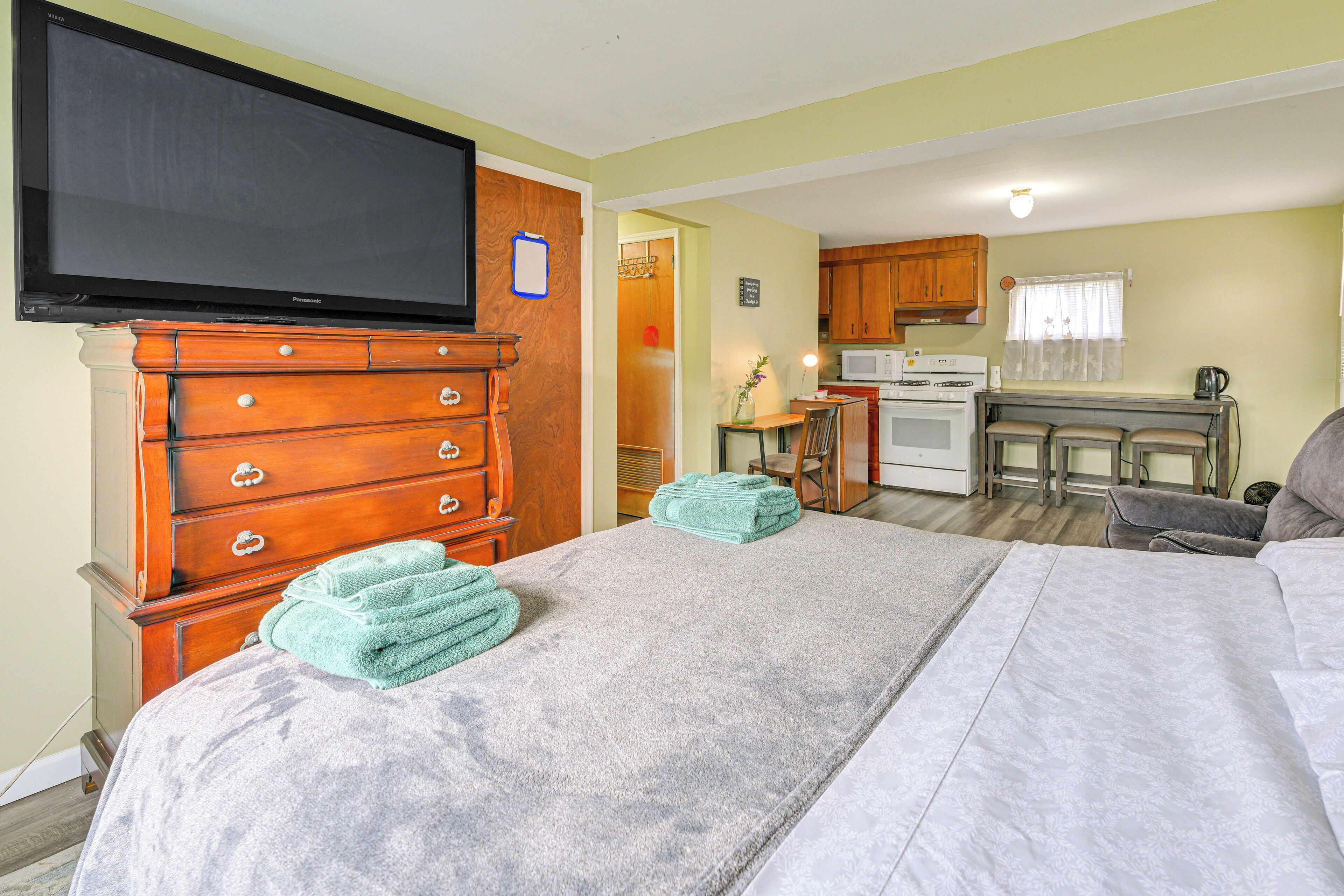A room with a bed, a TV, a wooden dresser, a kitchen area with a microwave, and a small table with a lamp.