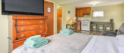 A room with a bed, a TV, a wooden dresser, a kitchen area with a microwave, and a small table with a lamp.