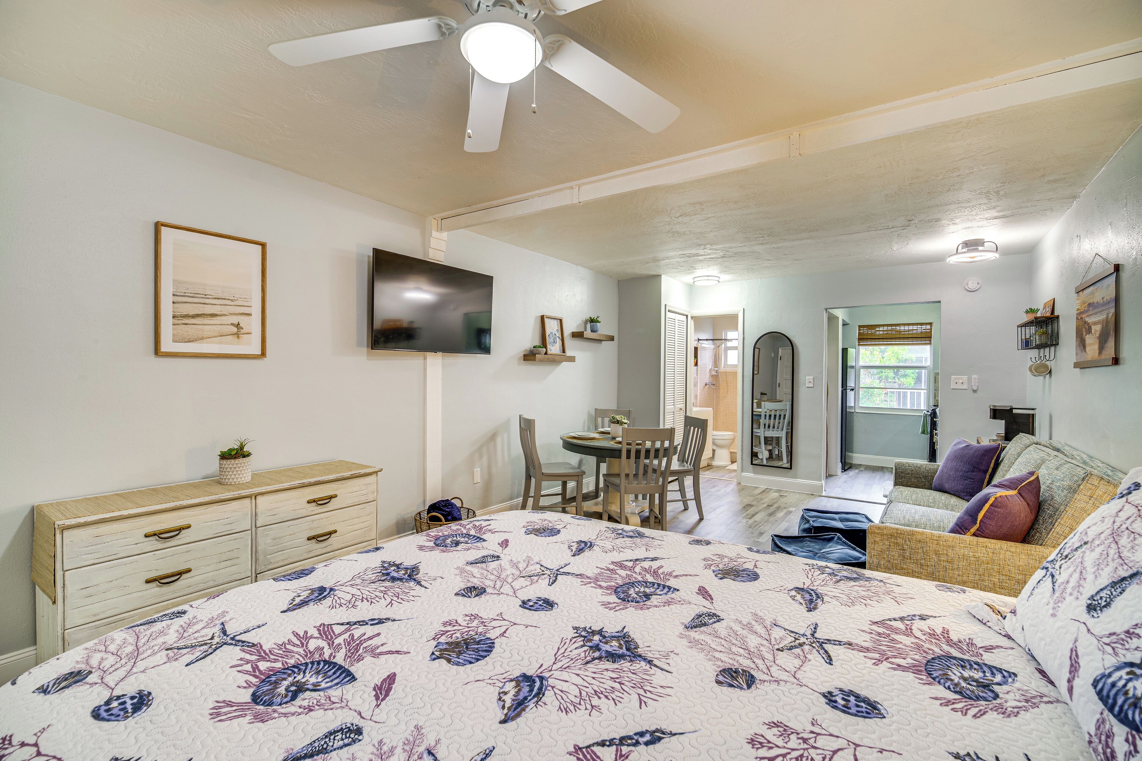 Un dormitorio con cama, cómoda, televisor, ventilador de techo y una zona de comedor con sillas y mesa.