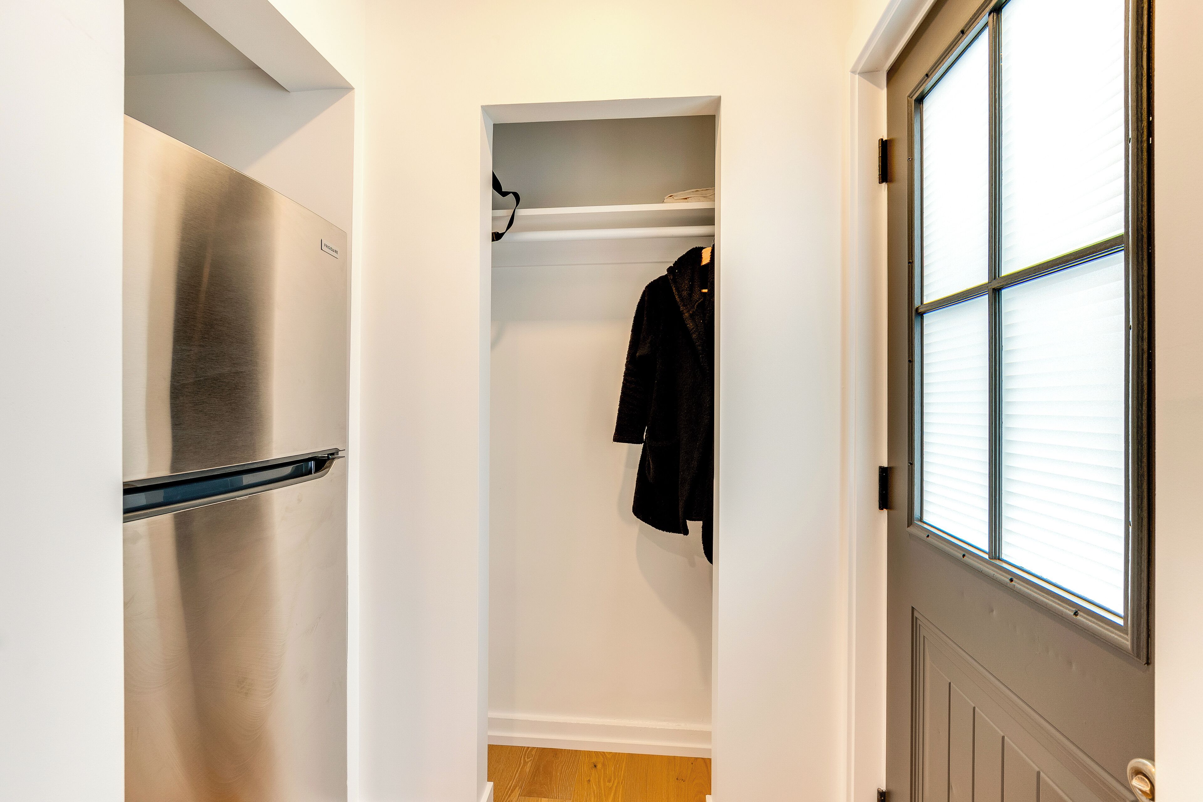 A narrow hallway with a stainless steel refrigerator on the left, a coat rack with a black jacket, and a window with blinds on the right.