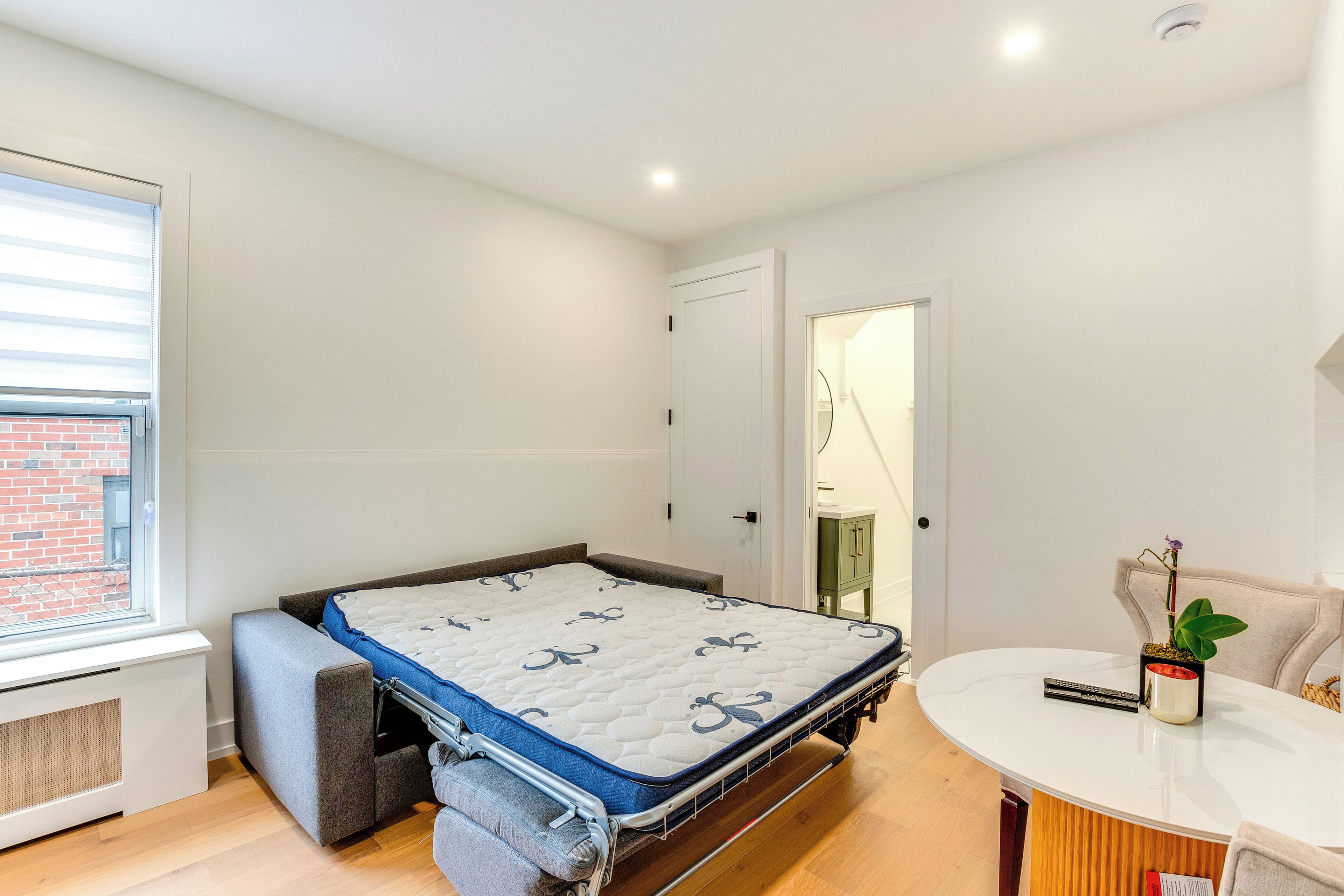 A compact bedroom with a pull-out bed, a small seating area, a white round table, and a window with blinds.