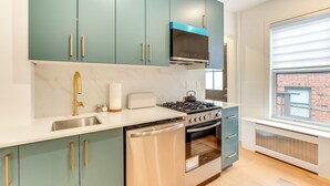 A modern kitchen with teal cabinets, stainless steel appliances, and a window with a view of a brick building. - Centrally Located & Pet Friendly: Yonkers Studio! (Yonkers)