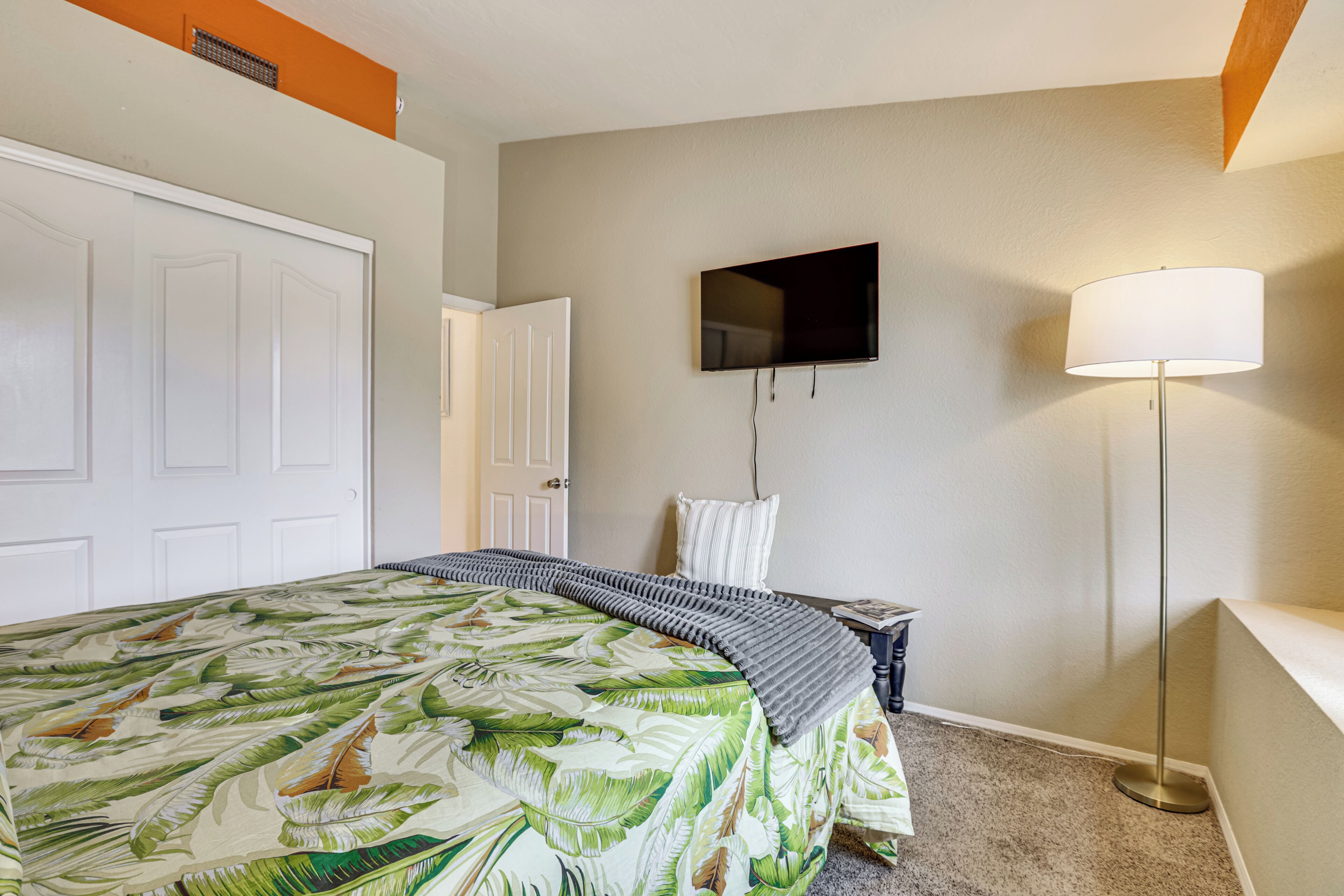 A bedroom with a bed, a television mounted on the wall, a floor lamp, and a door.