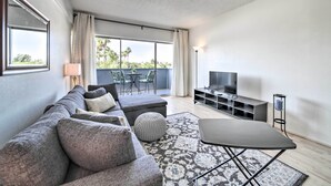 Un salon moderne avec un canapé gris, un pouf et une petite table. On y trouve un téléviseur posé sur un meuble, une grande fenêtre avec des rideaux et un balcon agrémenté de mobilier d’extérieur.