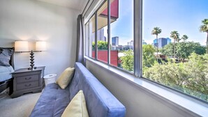 Une chambre avec une grande fenêtre, un lit, une table de chevet avec des lampes, et une vue sur un paysage urbain et des palmiers.