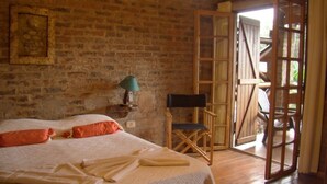 A rustic room with a brick wall, a bed with white linens, a wooden chair, and a large wooden door leading outside.