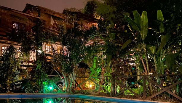 A nighttime pool area with a clear view of the pool's edge, surrounded by lush greenery and a building with warm lighting in the background.